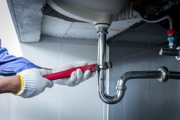Ein Klempner mit Handschuhen repariert mit einem Schraubenschlüssel ein Waschbeckenrohr in einem Badezimmer.